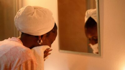 Woman with a towel wrapped around her hair drying her face in front of a mirror